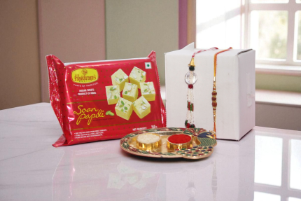 Bhaiya Bhabhi Rakhi gift combo featuring a designer Lumba Rakhi set, Haldiram Soan Papdi sweets, festive pooja thali with roli chawal, and white gift box – perfect Raksha Bandhan hamper for brother and sister-in-law. - YHOJA.COM